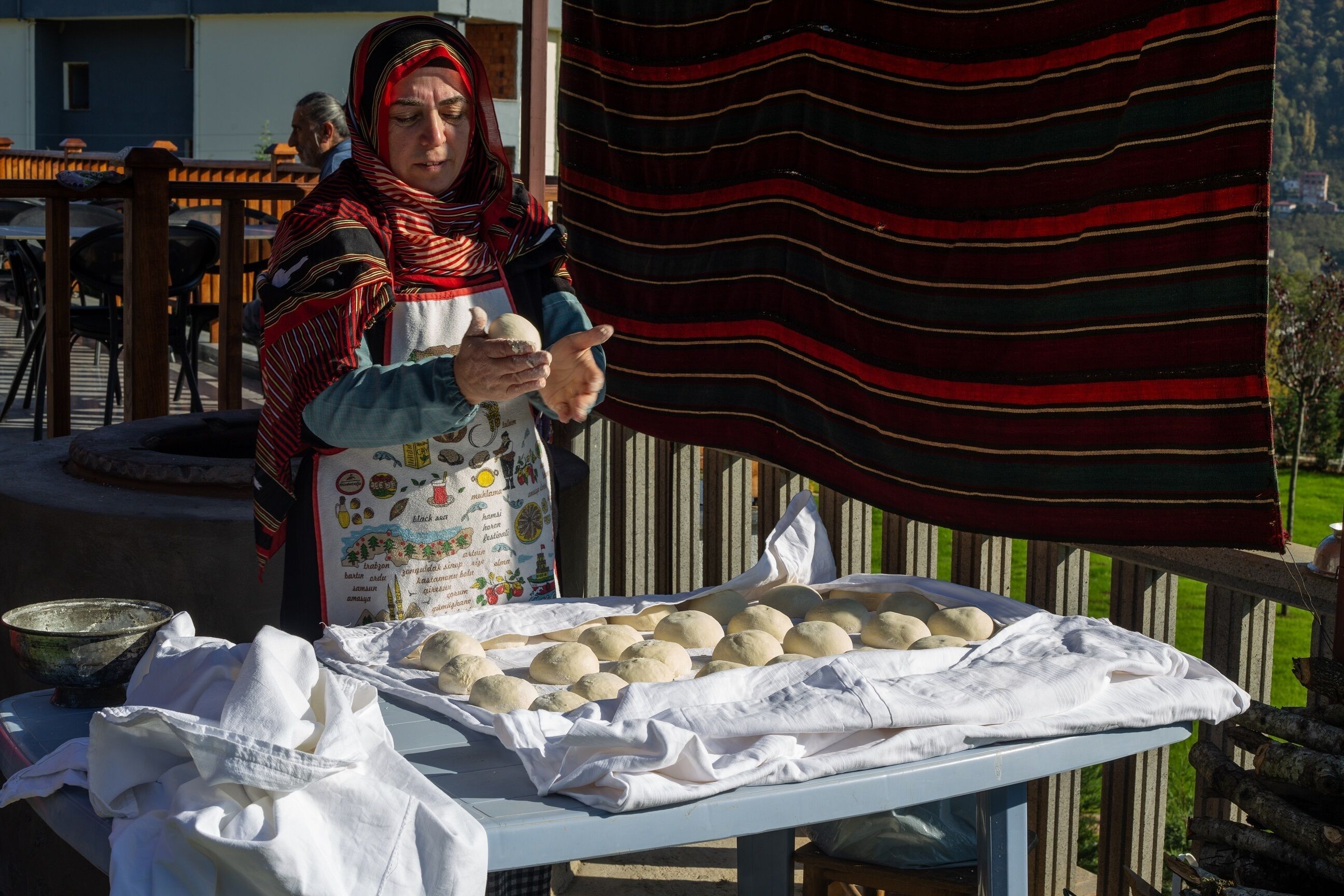 A person preparing food outdoors, with a table covered with cloths and a large striped fabric in the background.