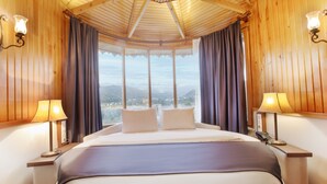 Quarto de hotel com uma cama grande, duas luminárias de cabeceira, um telefone e vista para as montanhas através de amplas janelas.