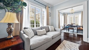 A living room with a grey sofa, a wooden side table with a lamp, a large plant, and a dining area with a chandelier. - Historic Retreat in Lake Claire (Atlanta)