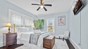 A bedroom with a bed, a nightstand, a lamp, a ceiling fan, a chair, and a wall-mounted TV. - Historic Retreat in Lake Claire (Atlanta)