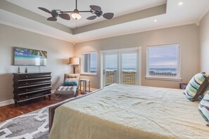 Un dormitorio con una cama grande, un aparador, una silla, una lámpara y vistas al océano a través de las ventanas.