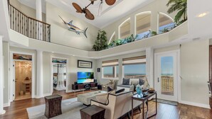 A spacious living area with a large TV, comfortable seating, wooden flooring, and a balcony with plants.
