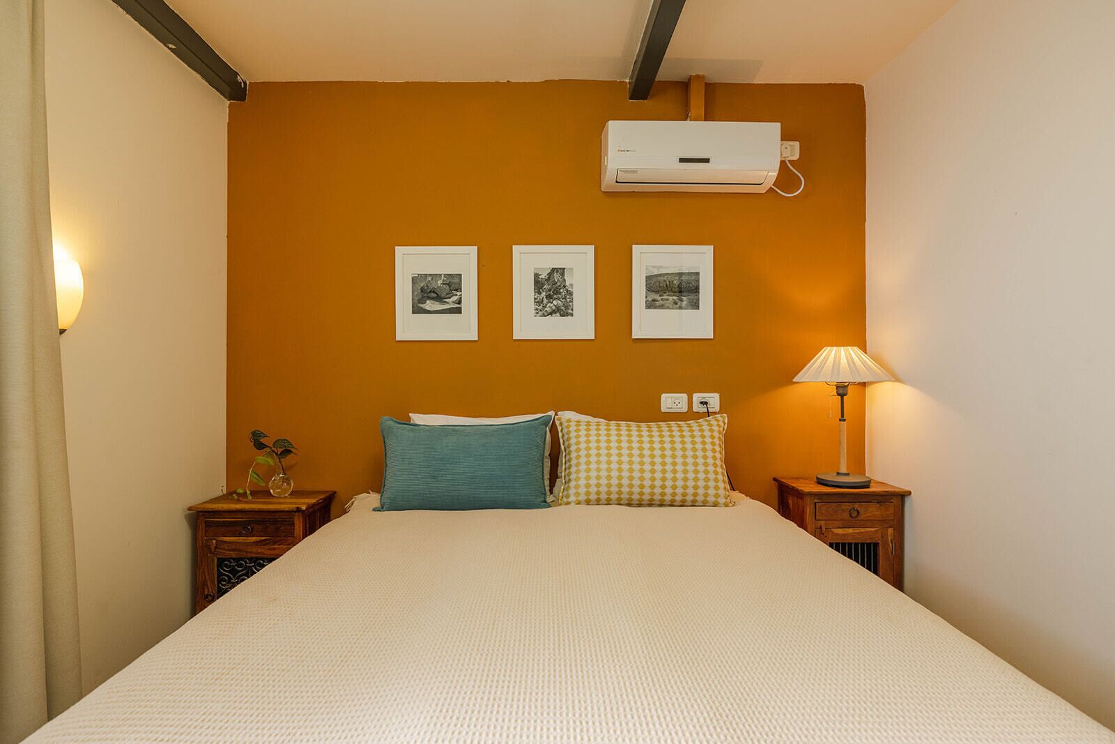 A bedroom with a bed, two pillows, a bedside table with a lamp, and a wall-mounted air conditioner.