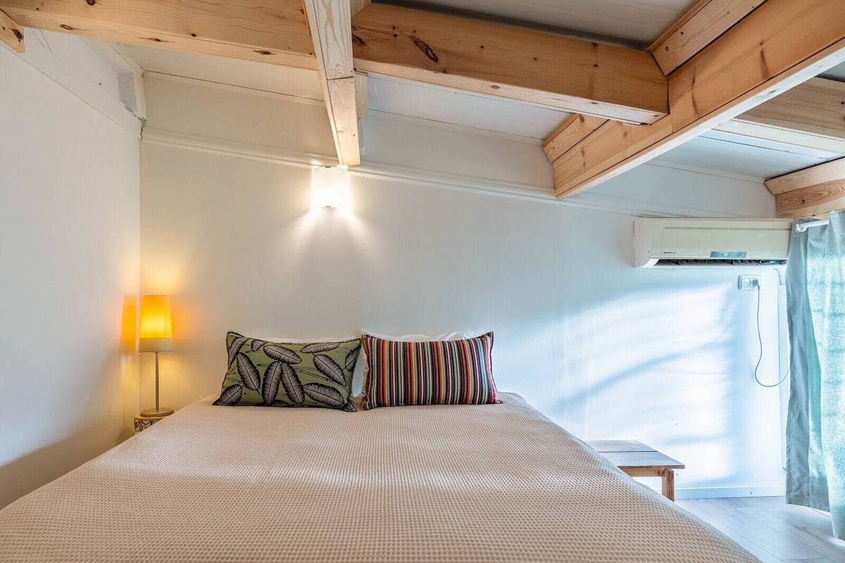 A bedroom with a bed, two pillows, a bedside table, a lamp, and an air conditioning unit.