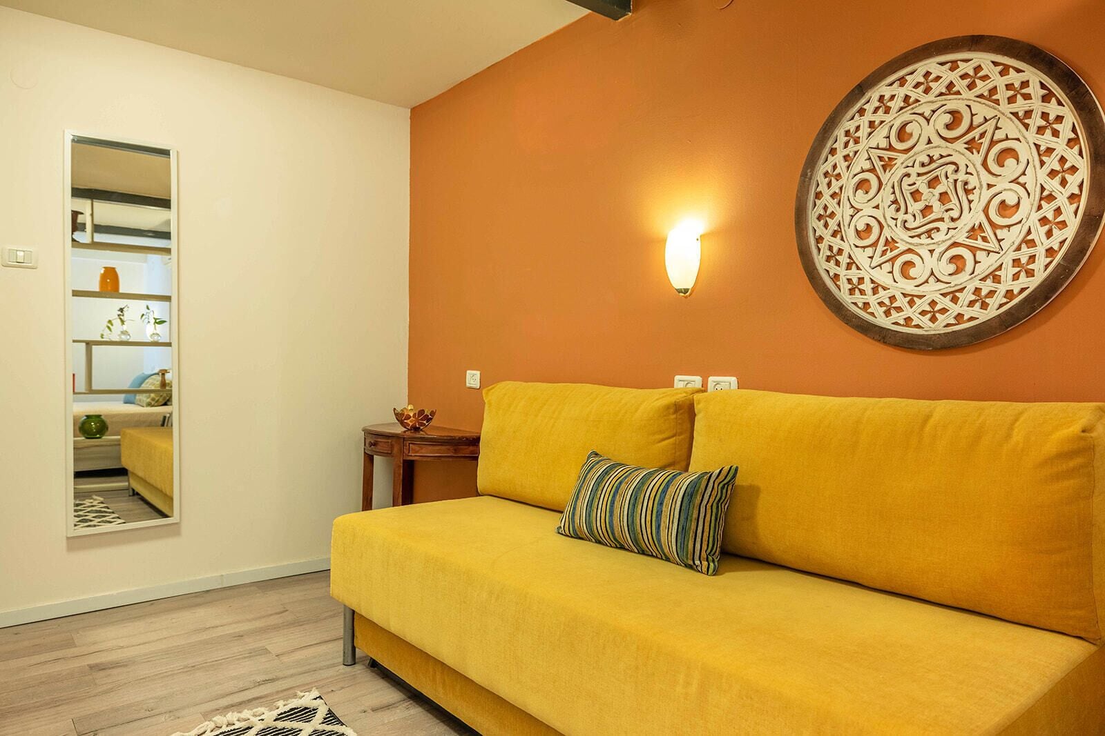 A yellow sofa with striped pillows, a decorative wall hanging, and a mirror reflecting a room with shelves and a vase.