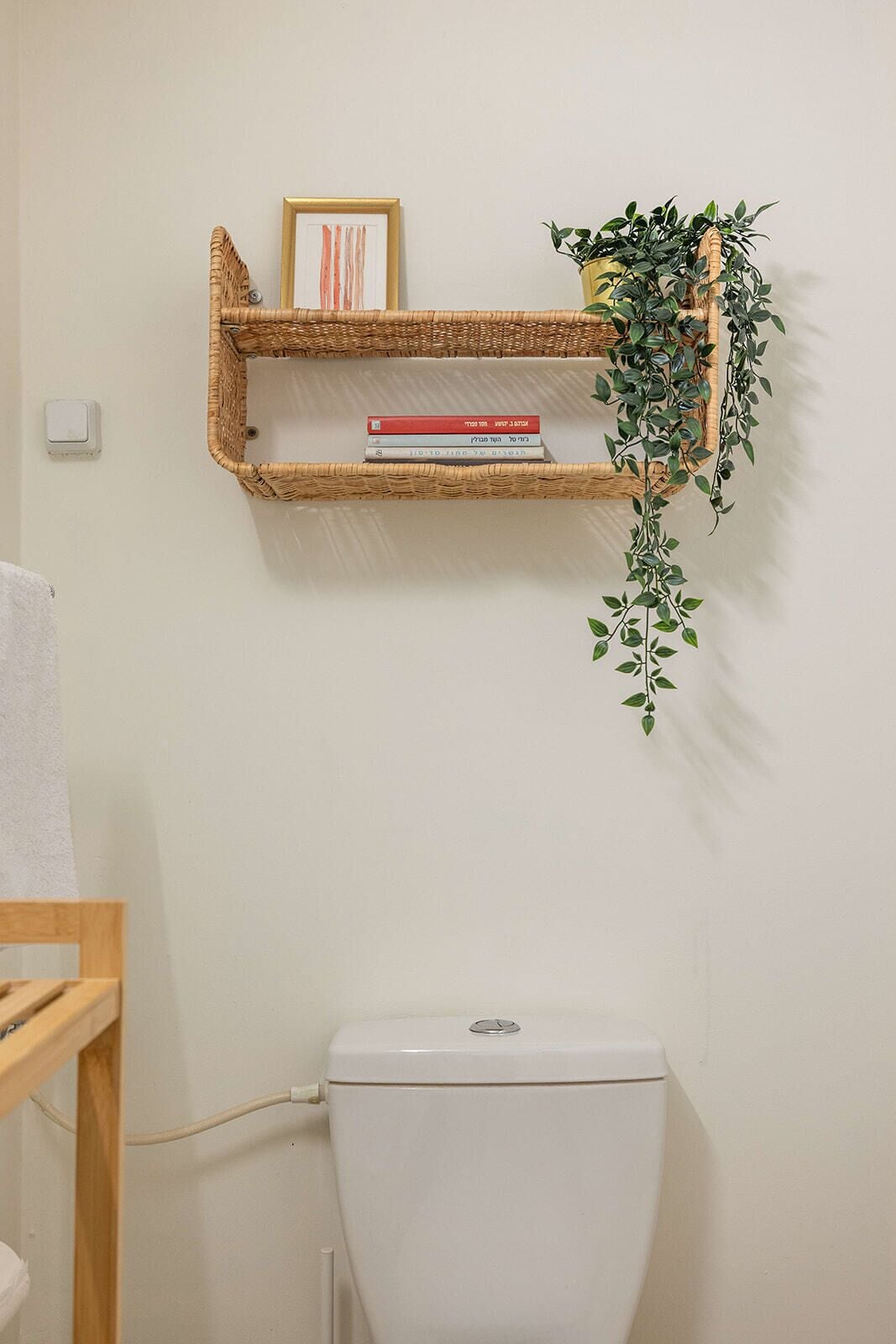 A bathroom with a white toilet, a wicker shelf with books and a plant, and a white towel on a rack.