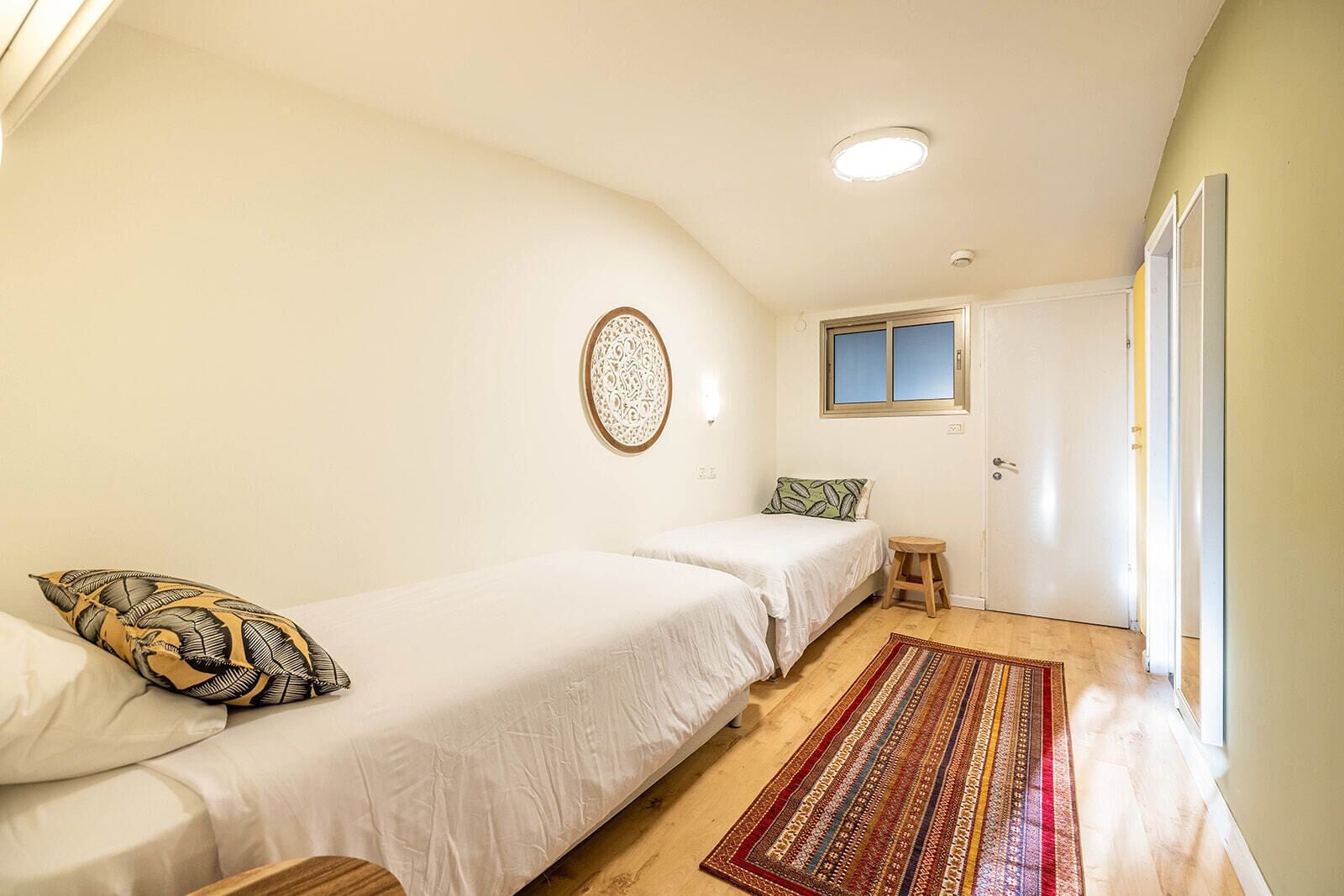 A bedroom with two single beds, a small stool, a patterned rug, and a window with a view.