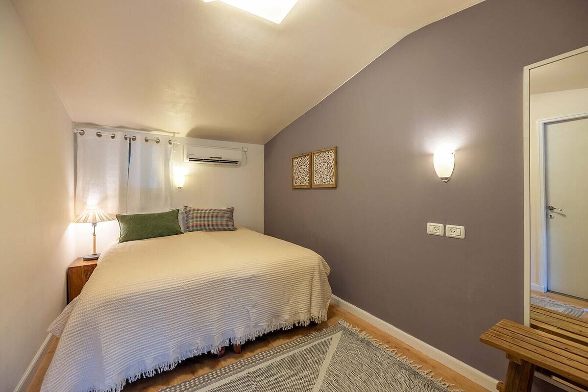 A bedroom with a bed, a nightstand, a lamp, a wall-mounted air conditioner, a door, and a small wooden stool.