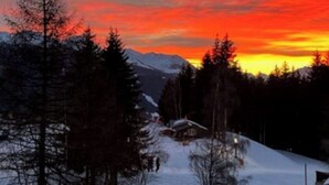 Eine verschneite Berglandschaft mit Sonnenuntergang, Bäumen und einer kleinen Hütte.