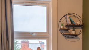 Een slaapkamer met een houten bed, een raam met uitzicht op een bakstenen gebouw, een klein zwart nachtkastje met een boek, en een wandplank met decoratieve objecten.
