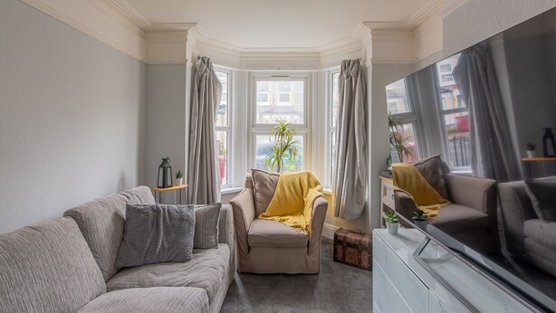 Een woonkamer met een bank, fauteuil en een televisie. Er is een raam met gordijnen en een plant op de vensterbank.