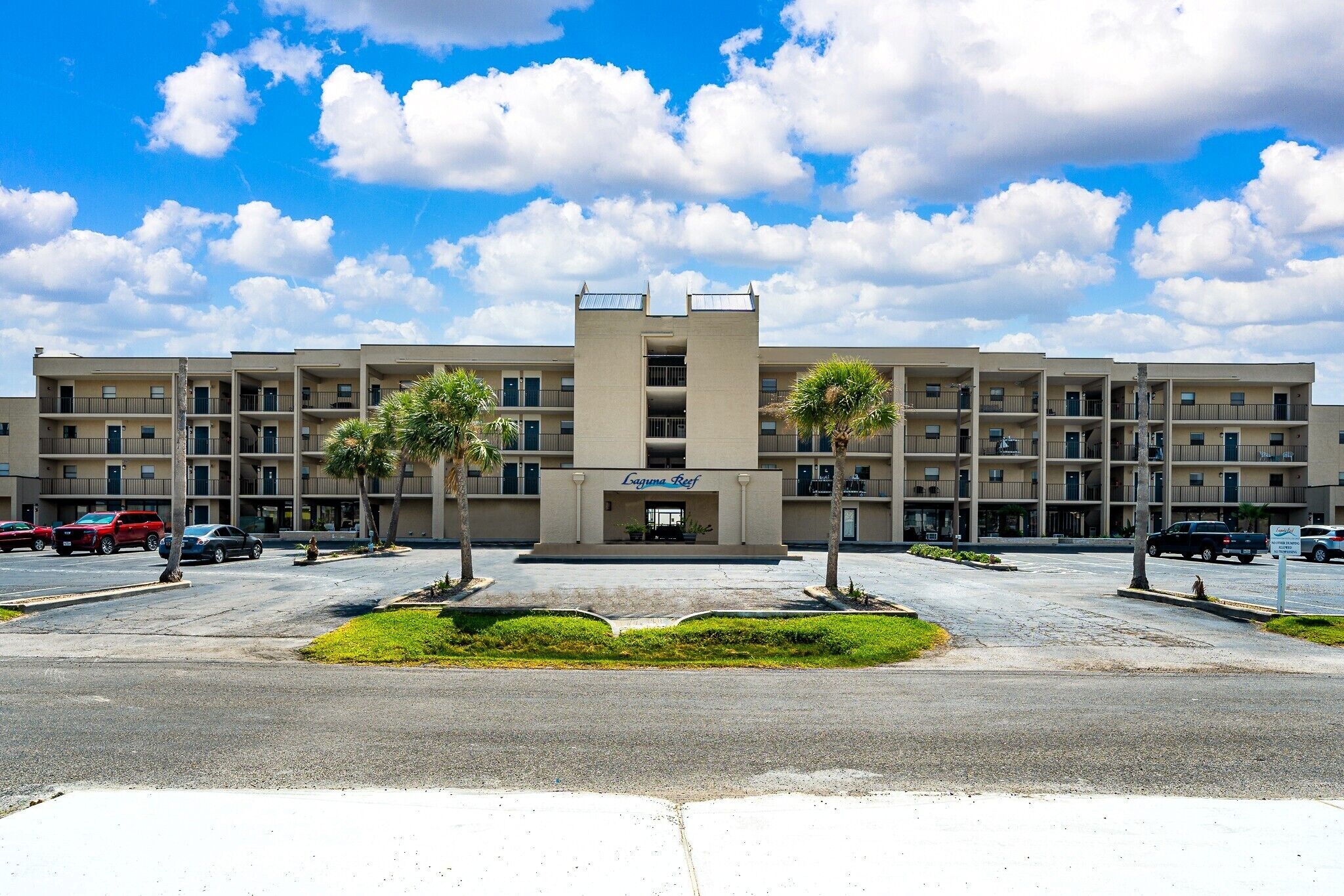 A multi-story hotel with a central entrance, parking lot, and palm trees.