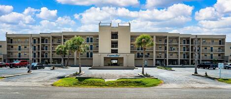 A multi-story hotel with a central entrance, parking lot, and palm trees.