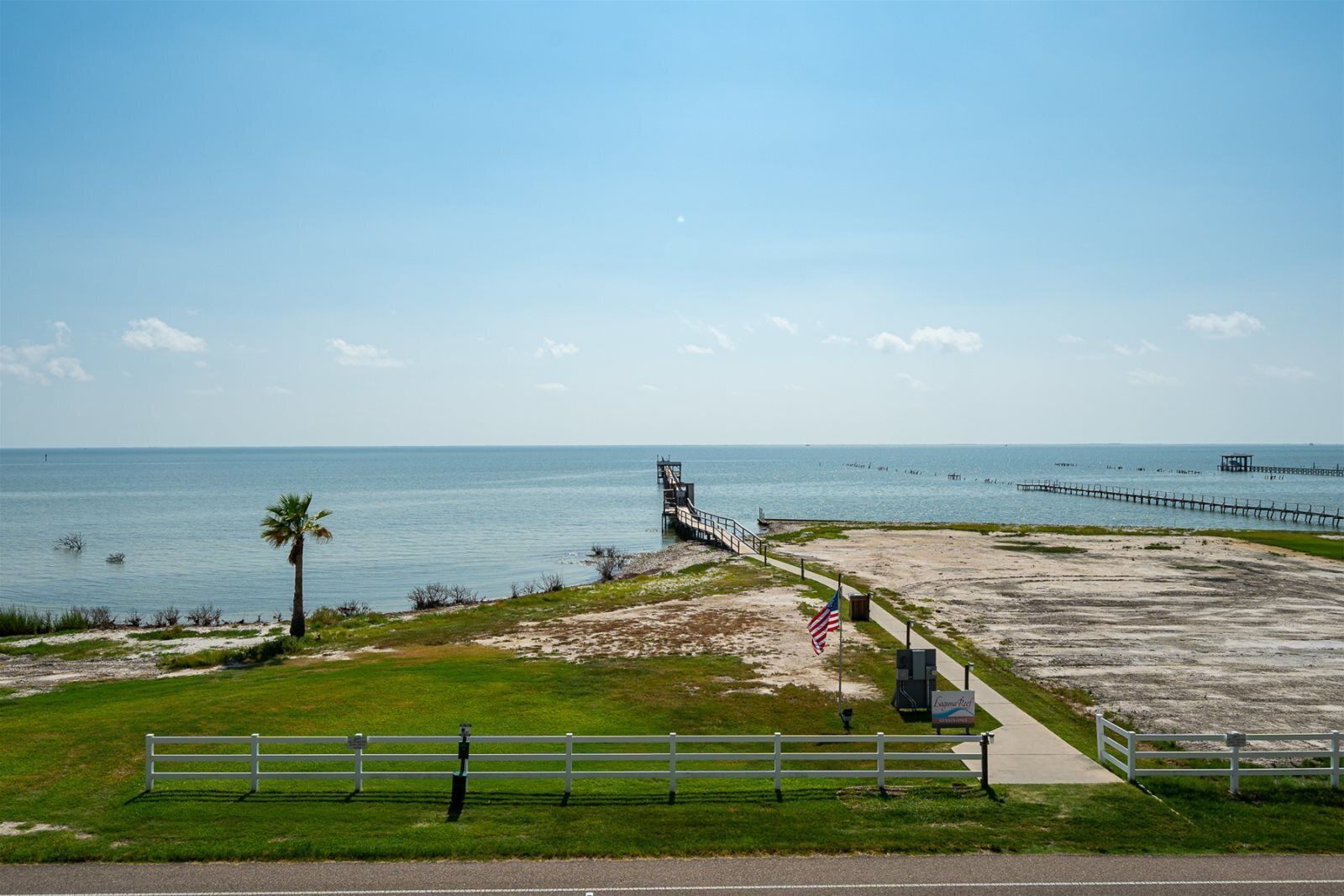 A beach with a wooden pier extending into the water, a palm tree, and a white fence along the shoreline.