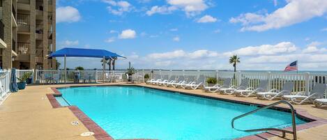A swimming pool with lounge chairs, a blue umbrella, and a view of the ocean.