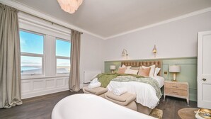 Un dormitorio con un ventanal grande, una cama con cabecera verde, una mesita de noche con lámparas y una bañera.
