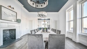 Comedor con una mesa larga, sillas grises tapizadas, chimenea y ventanas amplias.