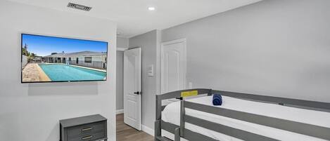 A bedroom with a bunk bed, a television mounted on the wall, a small bedside table, and a door leading to another room.