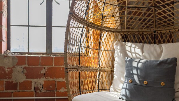 Um quarto com parede de tijolos, uma cadeira de vime e uma cama com almofadas brancas e cinzentas.