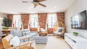 Uma sala de estar com parede de tijolos, ventilador de teto, televisão e um espaço de jantar com mesa e cadeiras.