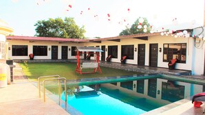 A swimming pool with a red swing chair, a small garden area, and a building with multiple windows and doors. - Lotus Resort (Chhatrapati Sambhajinagar)