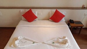 A neatly made bed with white linens and red pillows, a wooden nightstand with a phone and a lamp, and a wall socket. - Lotus Resort (Chhatrapati Sambhajinagar)