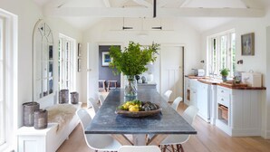 A spacious kitchen with a long table, white chairs, and a central island with a bowl of fruit and a vase of greenery. - Holmbush, Thornham, Norfolk (Thornham)