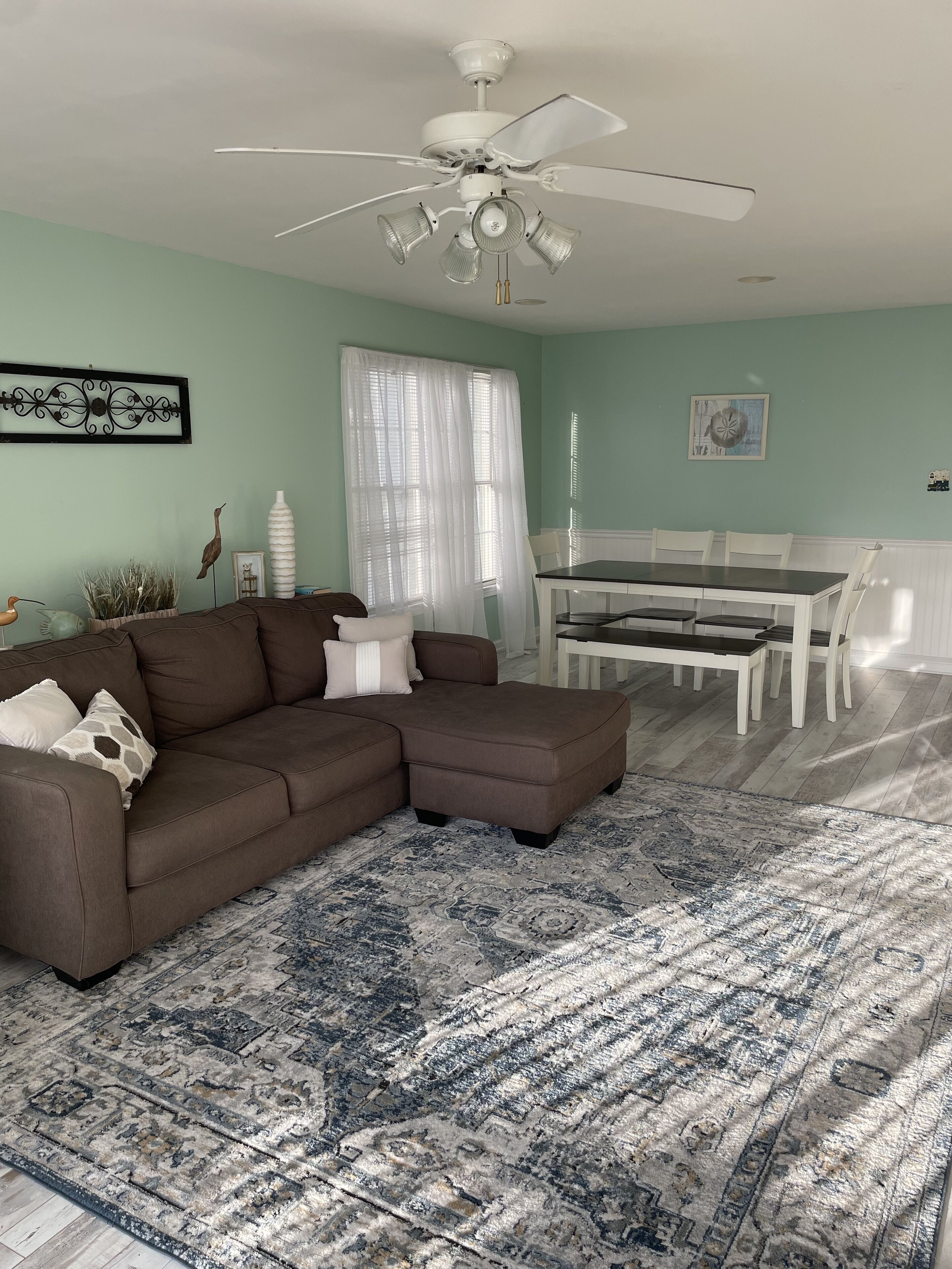 A living room with a brown sectional sofa, a dining table with chairs, a ceiling fan, and a patterned rug.