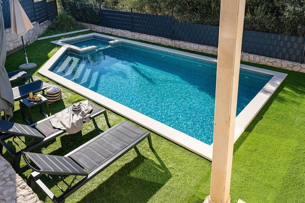 A rectangular swimming pool with a stone border, surrounded by artificial grass and equipped with lounge chairs and a table with a drink and snacks.
