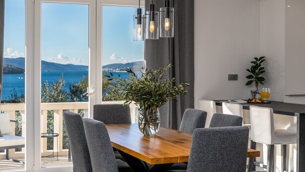 A modern dining area with a wooden table, grey upholstered chairs, and a vase with greenery. A kitchen area with a bar counter and a potted plant is visible in the background. - Villa Moreta with heated pool, Sauna, Hot-tub (Okrug Gornji)