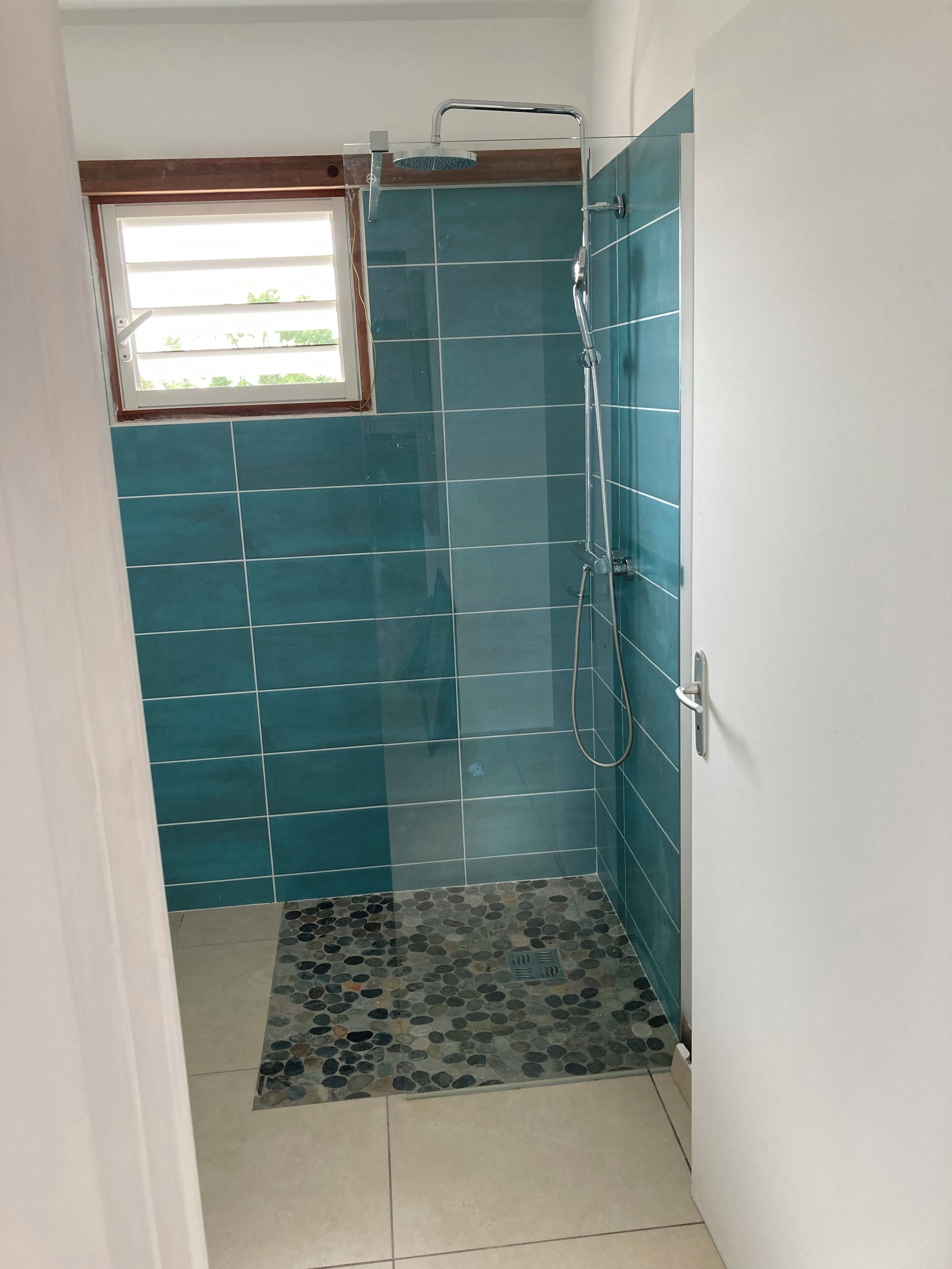 Un petit espace douche fermé, avec des carreaux de couleur turquoise et un sol à motifs de galets.