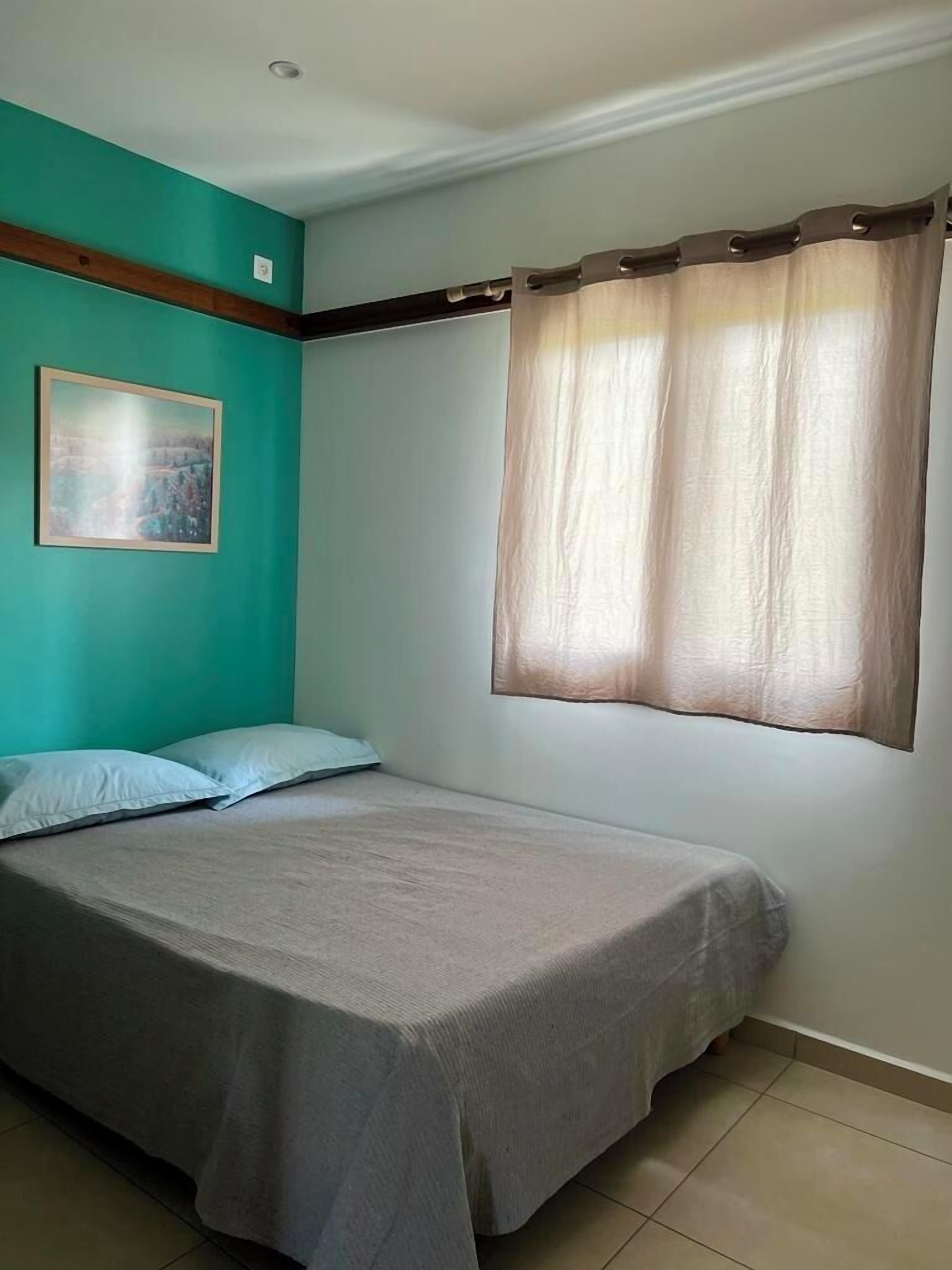 A single bed with a grey bedspread in a room with teal walls, a window with a brown curtain, and a framed picture on the wall.