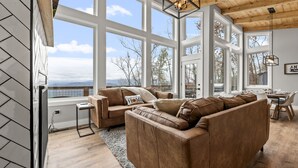 A spacious living room with a large brown leather sofa, a small side table, and a view of the outdoors through large windows. - New Modern Mountain Retreat with Spectacular Views (Sevierville)