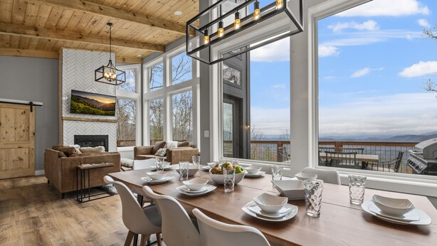 A modern dining area with a wooden table set for four, large windows offering a view of the outdoors, and a fireplace with a herringbone-patterned wall behind it. - New Modern Mountain Retreat with Spectacular Views (Sevierville)