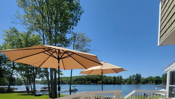 Une terrasse extérieure avec des meubles en plastique blanc et des parasols beige, donnant sur un lac où l’on peut voir des bateaux.