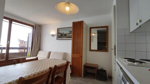 Ein Zimmer mit einem Bett, einem Esstisch, Stühlen, einem kleinen Hocker, einem Spiegel, einem Fenster und einer Küchenzeile.