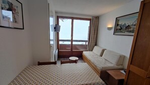 Ein kleines Zimmer mit einem Bett, einem Tisch und einem Stuhl. Es gibt ein Fenster mit Vorhängen und eine Tür.