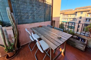 Une table en bois, entourée de chaises, se trouve sur un balcon dont le sol est recouvert de carreaux de terre cuite. On y aperçoit une plante en pot et, au loin, un immeuble d’habitation.
