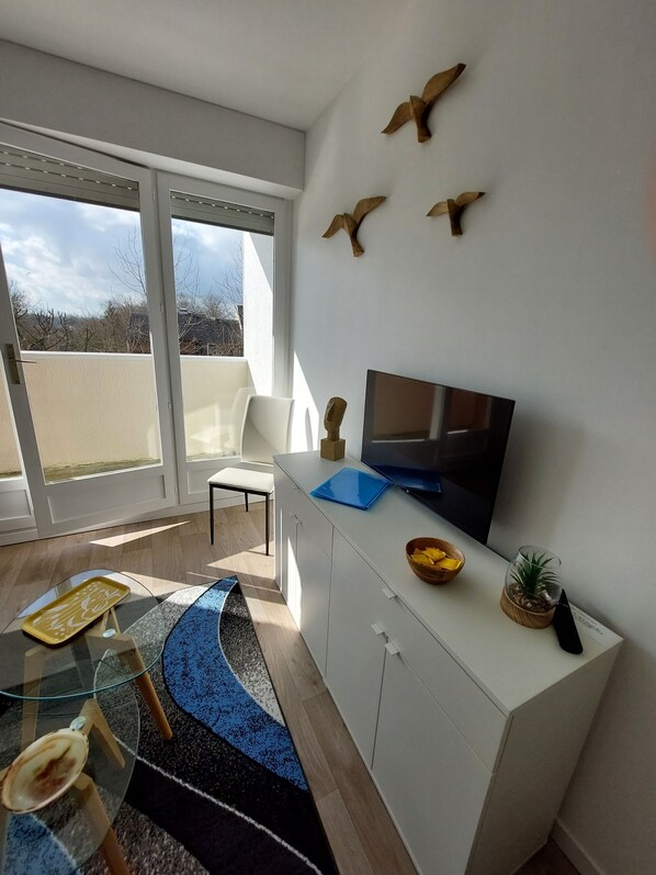 A modern living room with a large TV, white cabinets, a glass coffee table, a blue patterned rug, and a balcony with a view.