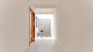 Un couloir avec une porte en bois, une petite table sur laquelle est posée une plante en pot, et une vue sur une pièce dotée d’une grande fenêtre.