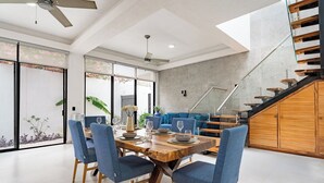 Un espace de repas moderne avec une table en bois et des chaises bleues, un escalier doté de rampes en verre et une vue sur un jardin, visible à travers de grandes portes coulissantes.