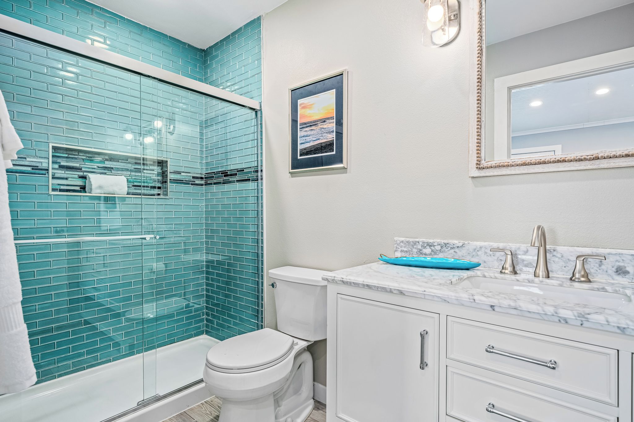A bathroom with a glass shower enclosure, a white toilet, a marble sink with a blue soap dish, and a framed picture on the wall.