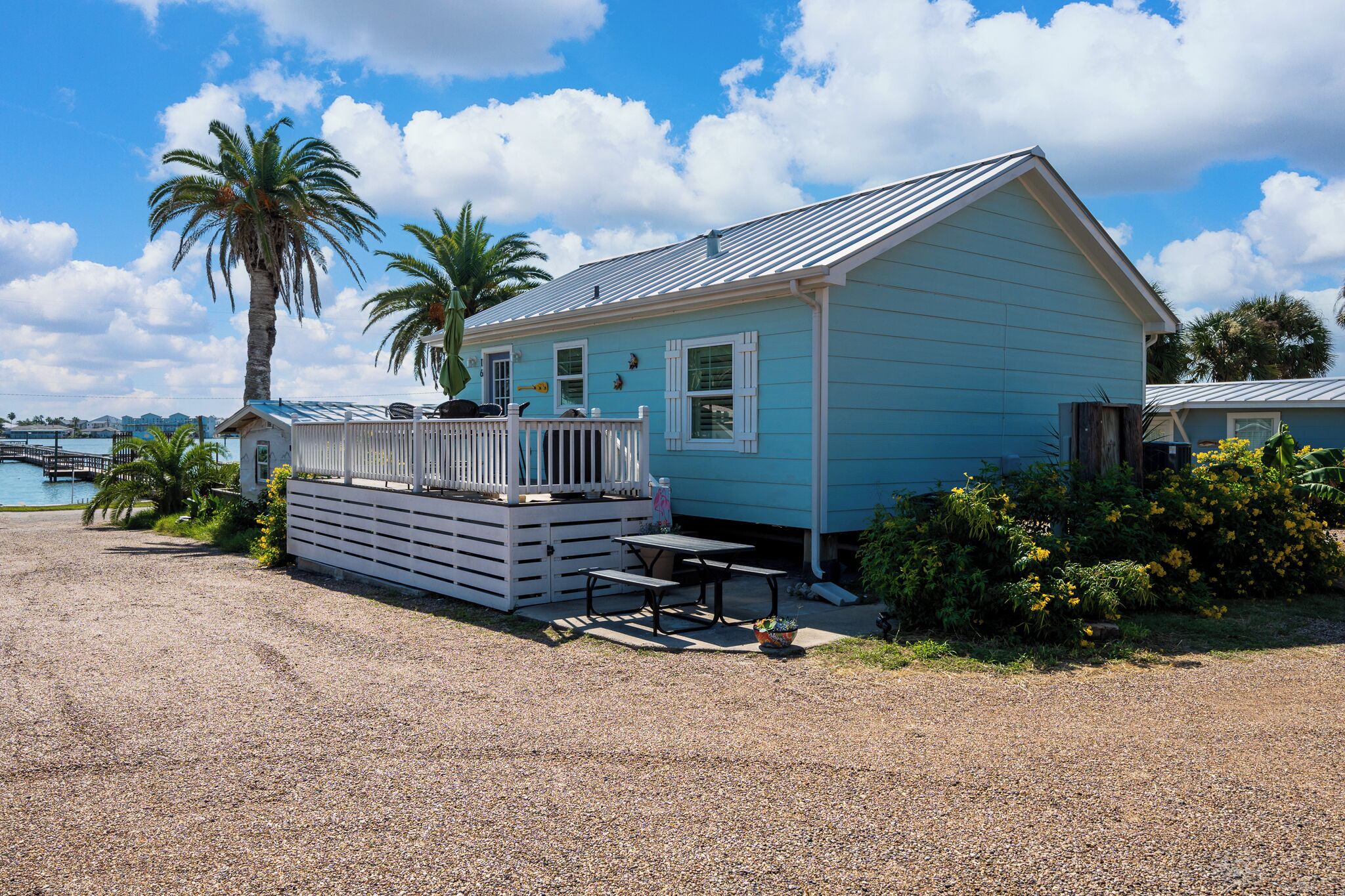 A small, light blue house with a porch and a table, surrounded by gravel and greenery.