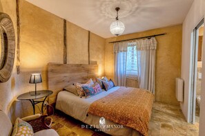 Un dormitorio con cabecera de madera, cama con ropa de cama estampada, mesita de noche con lámpara y una ventana grande con cortinas.