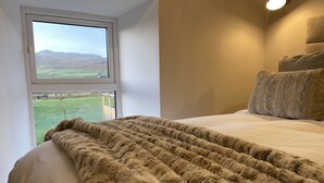 Un dormitorio con un ventanal grande, un edredón beige suave y una cama con dos almohadas.