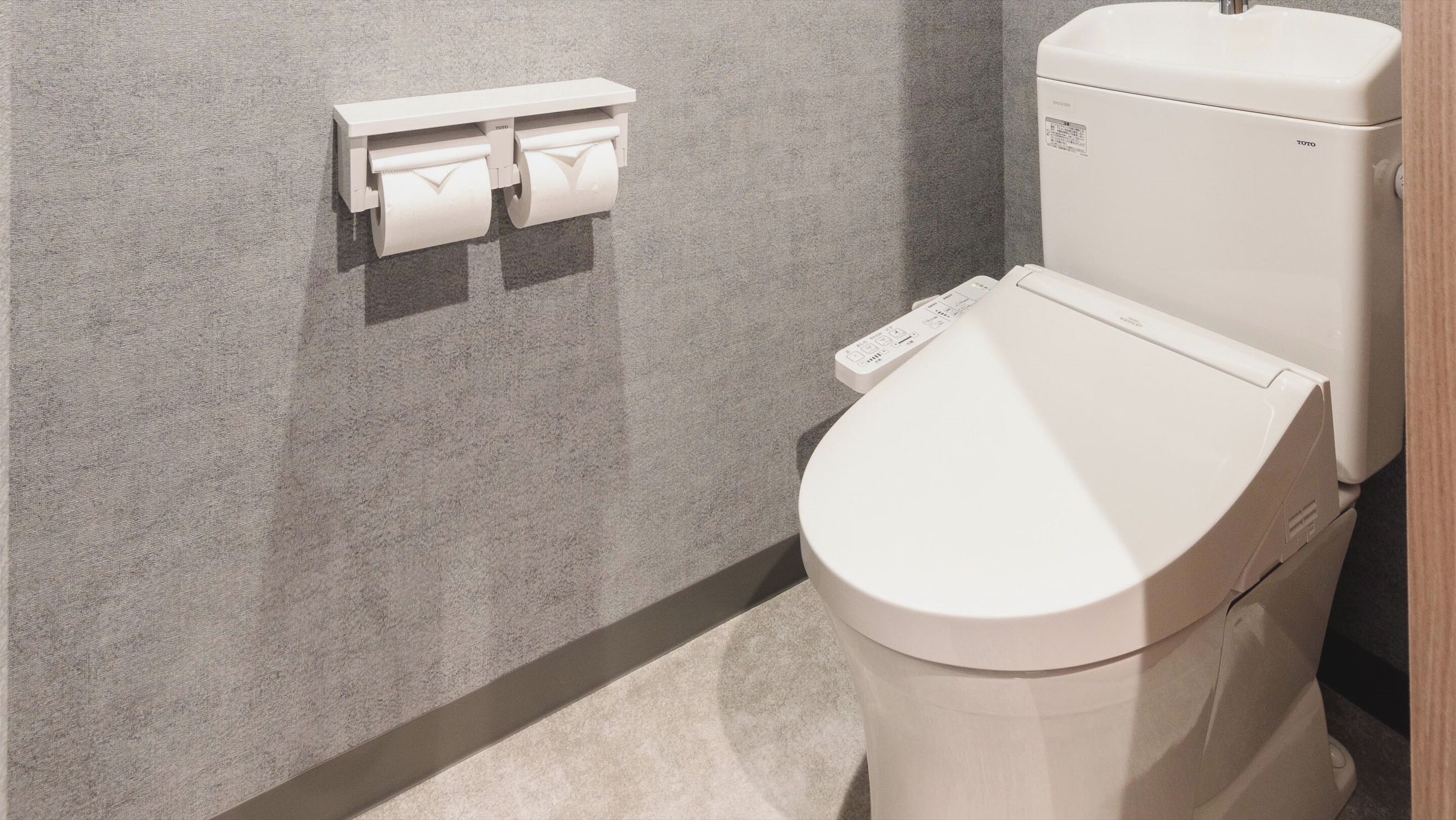 A modern toilet with a control panel, a wall-mounted toilet paper dispenser, and a plain wall.