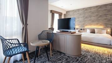 Habitación de hotel con cama, televisor sobre un mueble bajo, una mesita con silla y una ventana con cortinas.