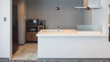 A modern kitchen with a white countertop, a built-in oven, a coffee maker, and a microwave. There are two pendant lights above the sink.
