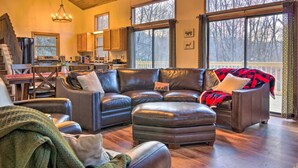 A spacious living room with a leather sectional sofa, a dining area with a wooden table and chairs, and large windows offering a view of the outdoors.