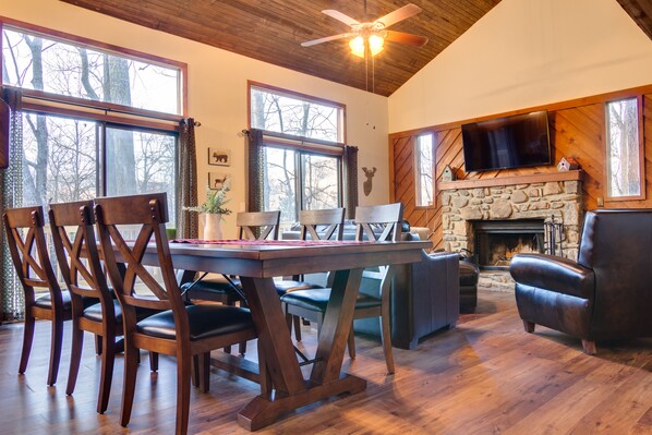 A cozy living room with a stone fireplace, wooden furniture, and large windows. - Brookhaven - Tranquil Maggie Valley Cabin with Deck & Fire Pit! (Maggie Valley)